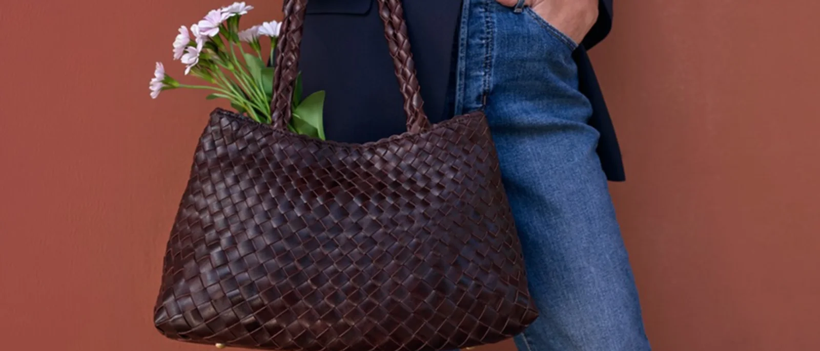 A woven leather tote with flowers in it, carried against a terracotta wall.